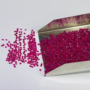May include: A metal scoop filled with loose pink ruby gemstones, with some spilling out onto a white surface.