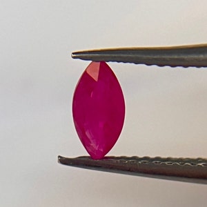 May include: A vibrant, deep red marquise-shaped gemstone held by tweezers. The gemstone is smooth and polished, with a brilliant shine.