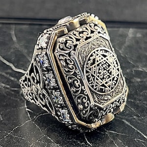 May include: A silver ring with a black enamel centre featuring a six-pointed star with Hebrew text. The ring is adorned with intricate filigree details and clear cubic zirconia stones.