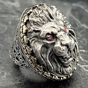 Puede incluir: Un anillo de plata con un diseño detallado de cabeza de león. Los ojos del león están engastados con piedras preciosas rojas. El anillo está rodeado por un borde dorado.