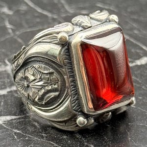 Könnte beinhalten: Ein silberner Ring mit einem verzierten Design und einem großen, rechteckigen, roten Edelstein. Der Ring hat detaillierte Schnitzereien auf dem Band und um die Edelsteinfassung, im Vintage- oder Antik-Stil.