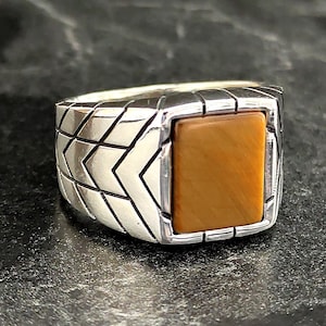 Könnte beinhalten: Ein silberner Ring mit einem quadratischen braunen Tigerauge-Edelstein, der in einem geometrischen Muster gefasst ist.