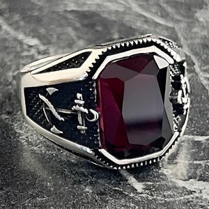 May include: A silver ring with a dark red gemstone and an anchor design.