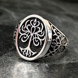 May include: A silver ring with an oval-shaped design featuring a stylized tree with intricate branches. The ring has a textured band with floral patterns.