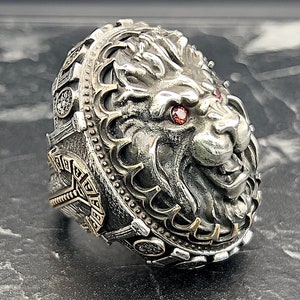 Può includere: Un anello d'argento con un designo dettagliato di testa di leone. Gli occhi del leone sono incastonati con pietre preziose rosse. L'anello è decorato con sculture intricate e una fascia dorata.
