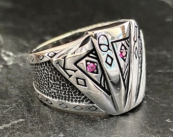 Puede incluir: Un anillo de plata con un diseño geométrico y dos piedras preciosas rosas. El anillo tiene una banda texturizada y una punta afilada en la parte superior.