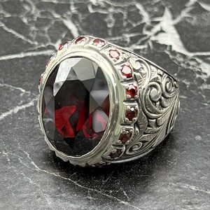 May include: A silver ring featuring a large, oval-cut, deep red gemstone. The ring is adorned with smaller red gemstones and detailed floral engravings on the band. The design showcases a classic, ornate style.
