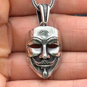 May include: A silver-colored pendant necklace featuring a detailed mask design. The mask has a stylized face with a prominent mustache and beard. The pendant hangs from a silver chain, creating a striking accessory.