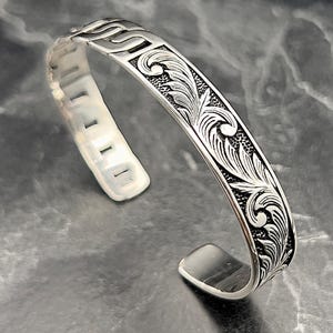 Könnte beinhalten: Silbernes Armreif-Armband mit einem komplizierten floralen Design. Das Armband ist offen und hat eine glatte, polierte Oberfläche.