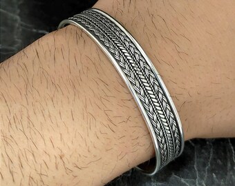 May include: A silver bracelet with a braided design. The bracelet is open and has a wide band.