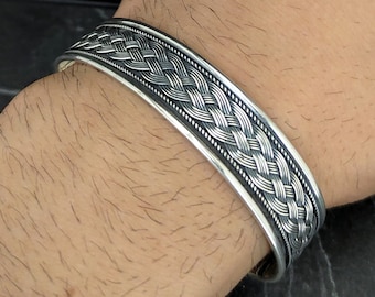May include: A silver bracelet with a braided design. The bracelet is wide and has a polished finish.