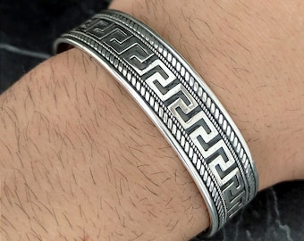May include: Silver bracelet with a Greek key pattern. The bracelet is wide and has a textured surface.