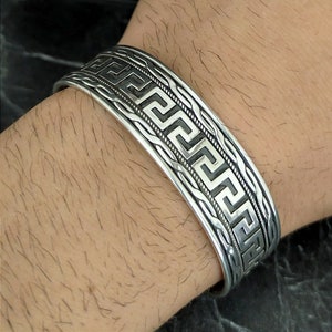 Puede incluir: Una pulsera de plata con un diseño de llave griega. La pulsera es ancha y tiene una superficie texturizada.