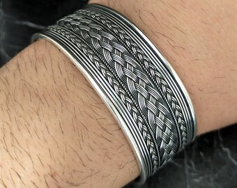 May include: A silver cuff bracelet with a braided design. The bracelet features multiple rows of woven patterns and is worn on a wrist. The metal has a polished finish, reflecting light. The overall style is bold and intricate.