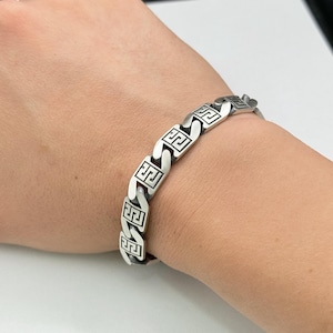 May include: A silver-toned metal chain link bracelet featuring a repeating black Greek key pattern engraved on each rectangular link. The bracelet has a polished finish and a secure interlocking design that wraps around the wrist.