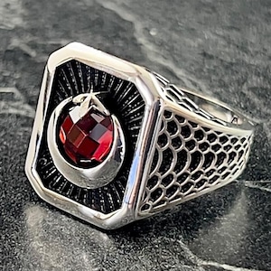 Könnte beinhalten: Silberner Ring mit rechteckiger Fassung, besetzt mit einem roten Edelstein in einem Halbmond-Design. Der Ring hat ein detailliertes, offenes Muster an den Seiten und eine polierte Oberfläche, die moderne und klassische Stile vereint.