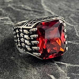 May include: A silver ring featuring a large, square-shaped red gemstone. The band of the ring has a detailed, textured design.