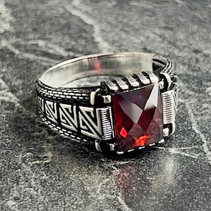 May include: A silver ring featuring a square-shaped red gemstone. The ring has a detailed design with intricate patterns.