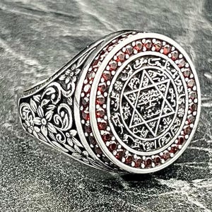 Może przedstawiać: Srebrny pierścionek ze szczegółowym kwiatowym wzorem na obrączce. Pierścionek ma okrągłą tarczę z motywem Gwiazdy Dawida i czerwonymi kamieniami szlachetnymi na obrzeżu. Pierścionek ma styl antyczny lub vintage.