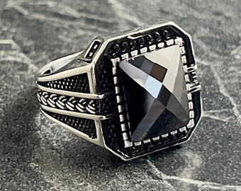 May include: A silver ring with a black square gemstone. The ring has a braided design on the band.