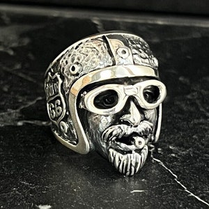 May include: A silver ring featuring a detailed sculpture of a man wearing a motorcycle helmet with goggles and a handlebar mustache. The helmet has a Route 66 sign on the side.