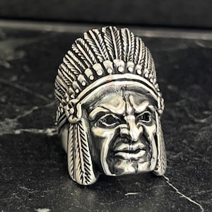 May include: A silver ring featuring a detailed sculpture of a Native American chief wearing a headdress. The chief's face is stern and the headdress is adorned with feathers.