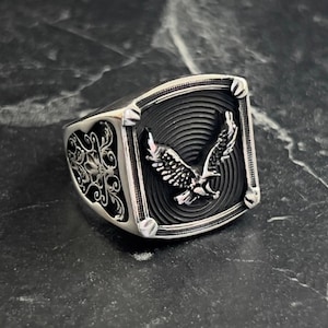 Könnte beinhalten: Ein silberner Ring mit einem quadratischen Design, das einen fliegenden Adler zeigt. Der Adler ist mit aufwendigen Schnitzereien verziert und vor einem schwarzen Hintergrund platziert. Der Ringband ist mit floralen Mustern verziert.