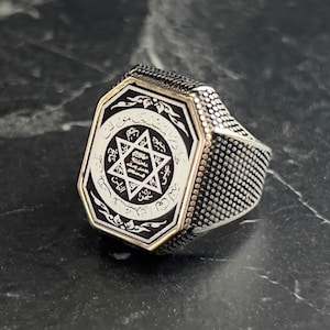 May include: A silver ring with a black octagonal stone featuring a white star of David and Arabic script.