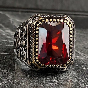 Könnte beinhalten: Ein silberner Ring mit einem verzierten Design und einem großen, rechteckigen roten Edelstein.