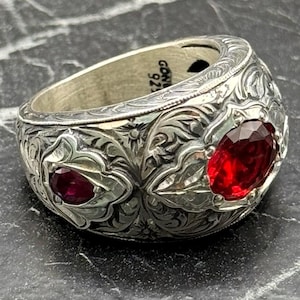 May include: A silver ring with ornate floral engravings and two oval-cut red gemstones. The ring is marked "GDN 925" on the inside. The ring has a wide band and a detailed, textured surface.