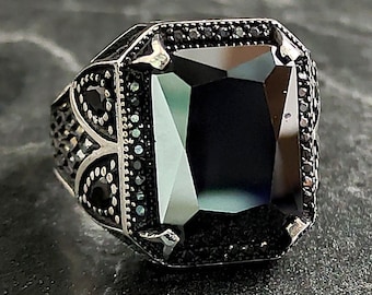 May include: A silver ring with a large, black, square-shaped gemstone. The ring is decorated with smaller black gemstones around the edge of the setting.