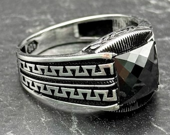 May include: A silver ring with a large, square, black gemstone. The ring has a detailed design around the band with a geometric pattern. The ring is displayed on a dark, textured surface.