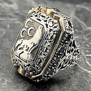 Op de afbeelding: Een zilveren ring met een sierlijk ontwerp met een wolf die huilt naar de maan. De ring heeft een zwarte steen in het midden en een gouden band.
