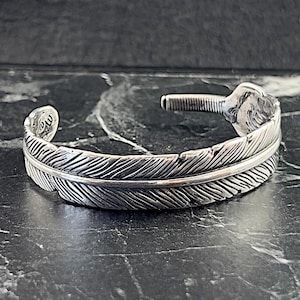 May include: A silver feather cuff bracelet with a textured design. The bracelet is open and has a spring clasp.