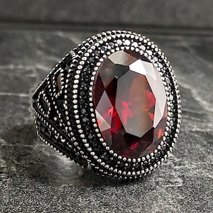 Puede incluir: Un anillo de plata con una gran piedra preciosa roja facetada rodeada por un borde de piedras negras.