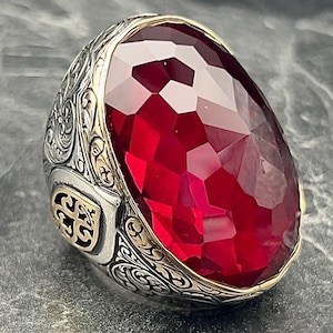 Può includere: Un anello d'argento con un designo ornato e una grande pietra rossa sfaccettata.