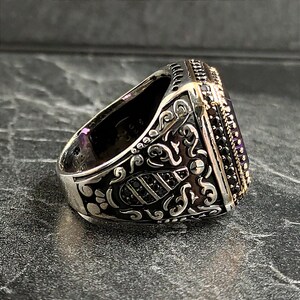 May include: A silver ring with an ornate design and a large, square, purple gemstone. The ring is set with smaller black gemstones around the edge of the stone.