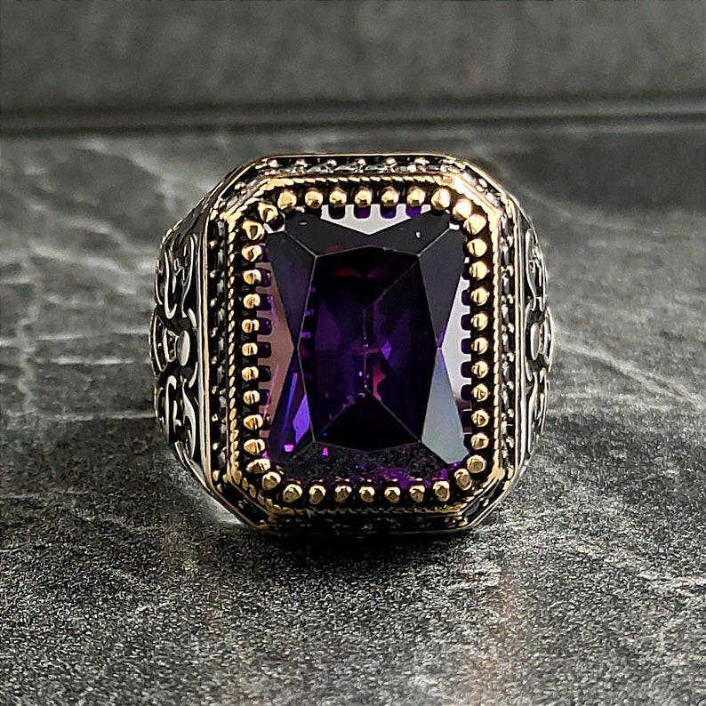 May include: A silver ring featuring a large, faceted, purple gemstone set in a gold-toned bezel. The ring has intricate, ornate details around the gemstone.