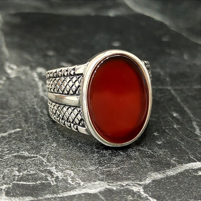 May include: A silver ring with a red carnelian stone set in a bezel setting. The band of the ring has a textured design.