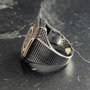 May include: A silver ring with a square face featuring a detailed design. The band of the ring has a textured finish.