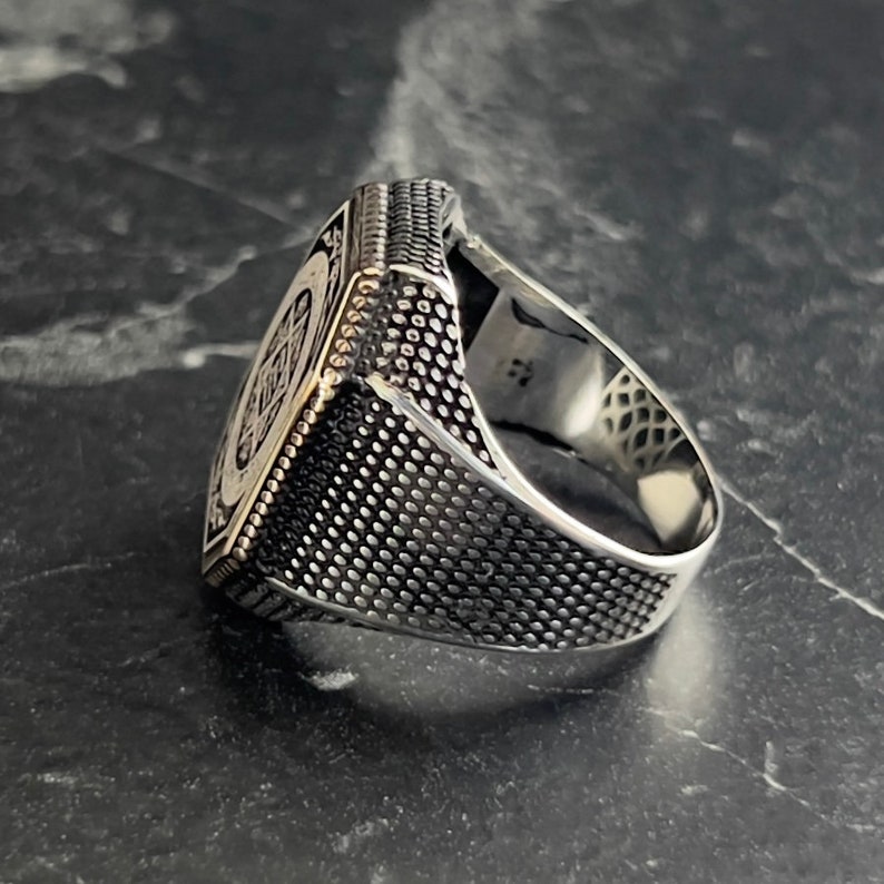 May include: A silver ring with a square face featuring a detailed design. The band of the ring has a textured finish.
