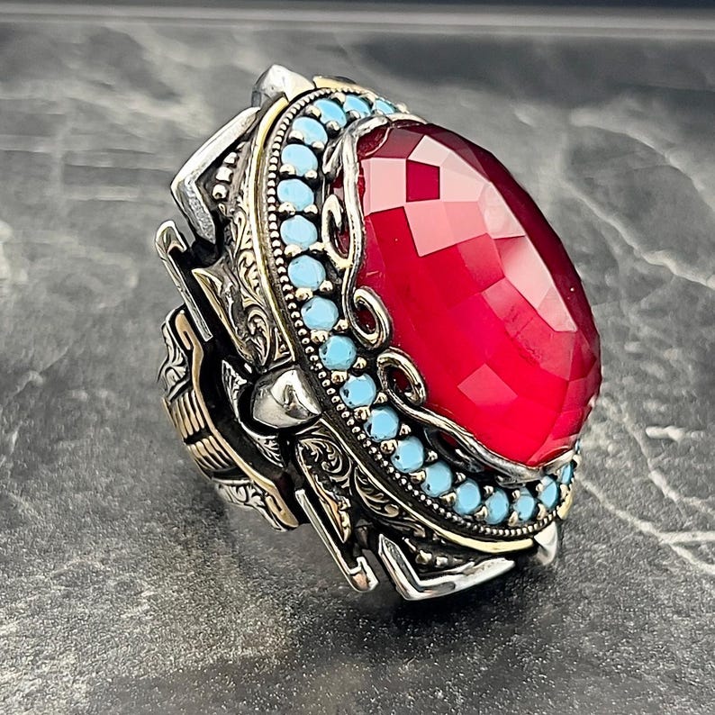 May include: A silver ring featuring a large red gemstone set within a border of turquoise stones. The ring is intricately detailed and has a gold accent.