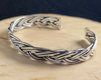 May include: A silver braided cuff bracelet. The bracelet is made of multiple strands of silver wire that are woven together to create a braided design.