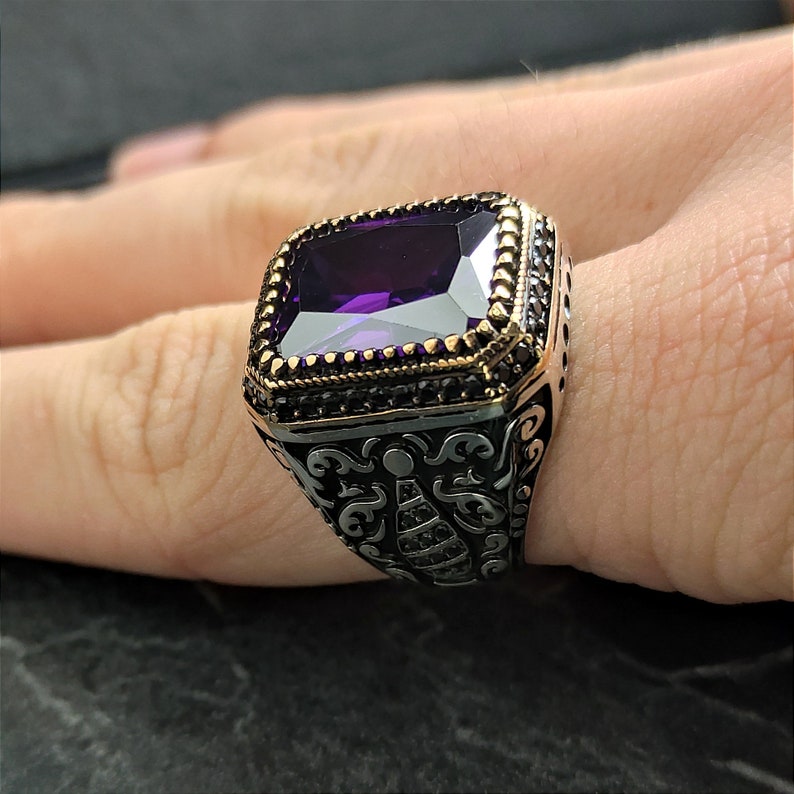 May include: A silver ring featuring a large, rectangular, purple gemstone set in a gold bezel. The ring has a black oxidised finish and intricate floral designs.