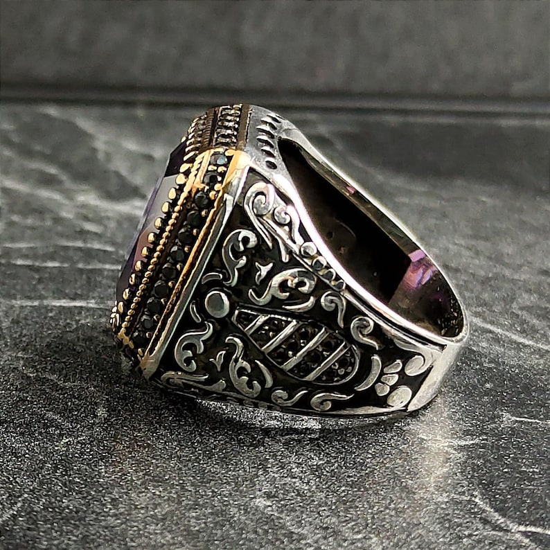 May include: A silver ring with an ornate design and a large, square, purple gemstone. The ring is set with small black stones around the gemstone.