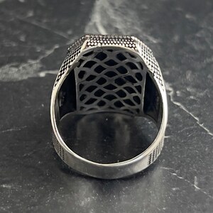 May include: Silver ring with a detailed, intricate design. The ring has a square-shaped band with a textured surface.