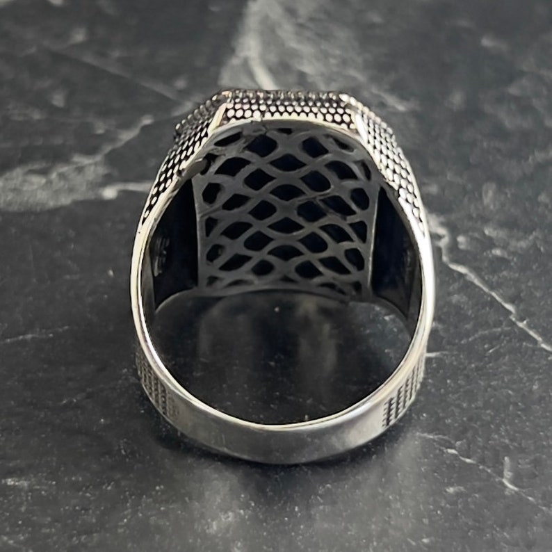 May include: Silver ring with a detailed, intricate design. The ring has a square-shaped band with a textured surface.