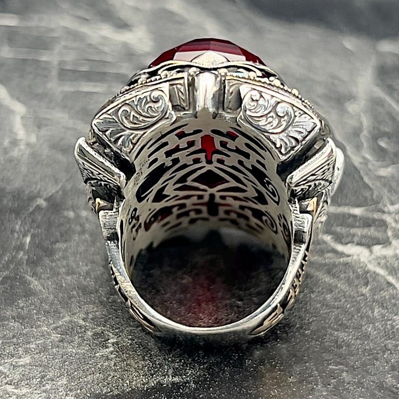May include: A silver ring featuring an elaborate design and a large red gemstone. The ring is set with a single, large, red gemstone in the centre. The band of the ring is adorned with a detailed, intricate design.