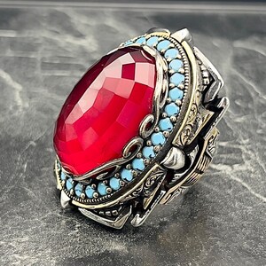May include: A silver ring featuring a large, faceted, red gemstone surrounded by smaller, turquoise gemstones. The ring has intricate, detailed designs and a gold accent.