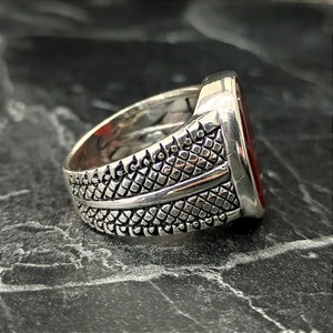 May include: A silver ring with a red stone set in a square shape. The band of the ring has a textured design.